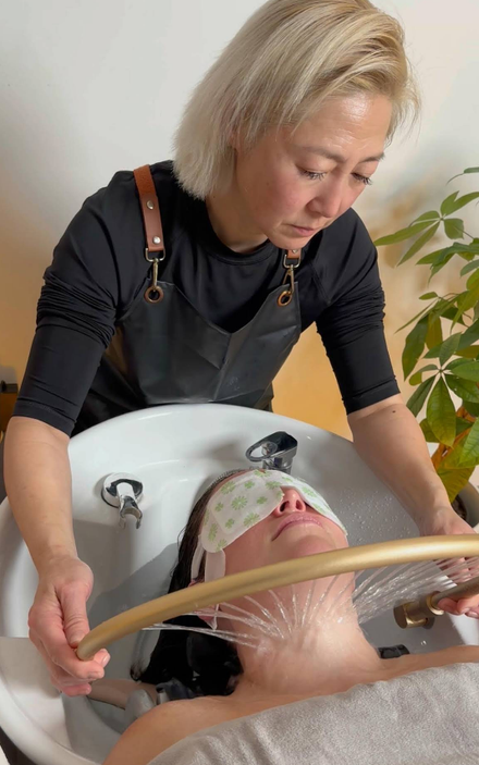 A stylist washes a client's hair in a salon. The client wears an eye mask and the stylist holds a showerhead.