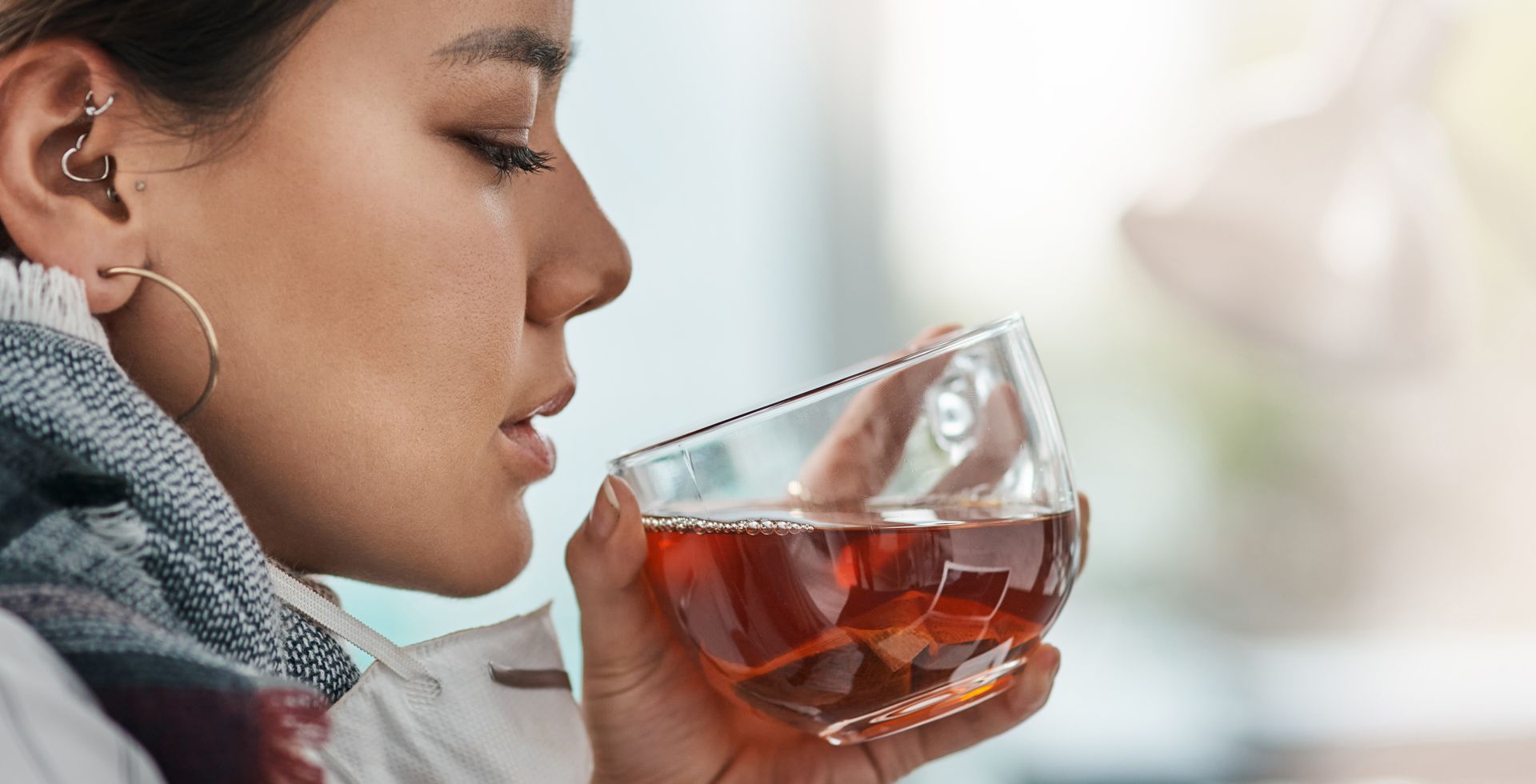 Woman sipping tea, eyes closed, enjoying the aroma. Clear glass, warm liquid, indoor setting.