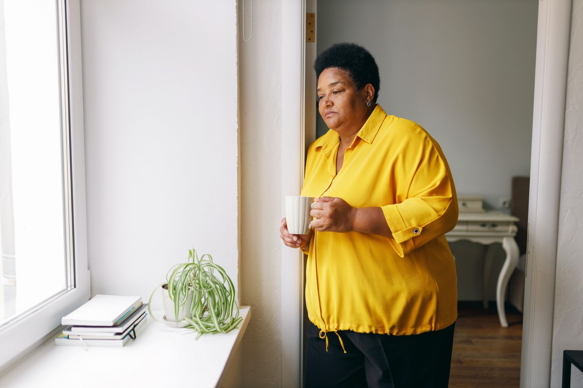 Woman in yellow shirt looking out window, holding mug. White window frame, plant, books.