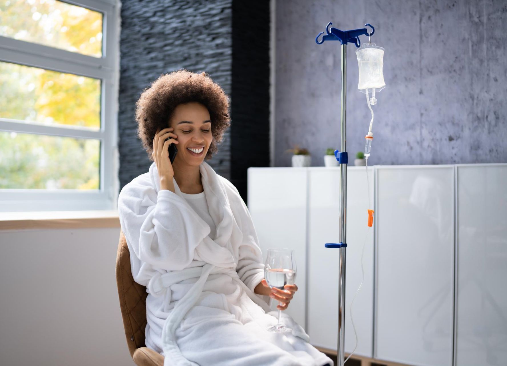 A woman in a bathrobe is sitting in a chair talking on a cell phone while receiving IV therapy infusion.