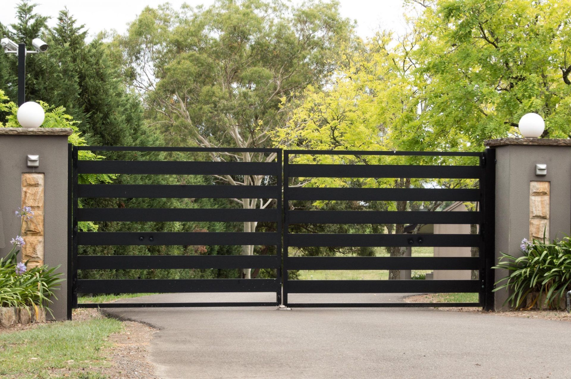 A black gate is open to a driveway with trees in the background