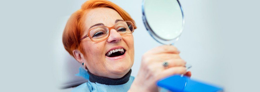 Older woman looking at her teeth on a mirror. Difference between implant dentures and regular dentures