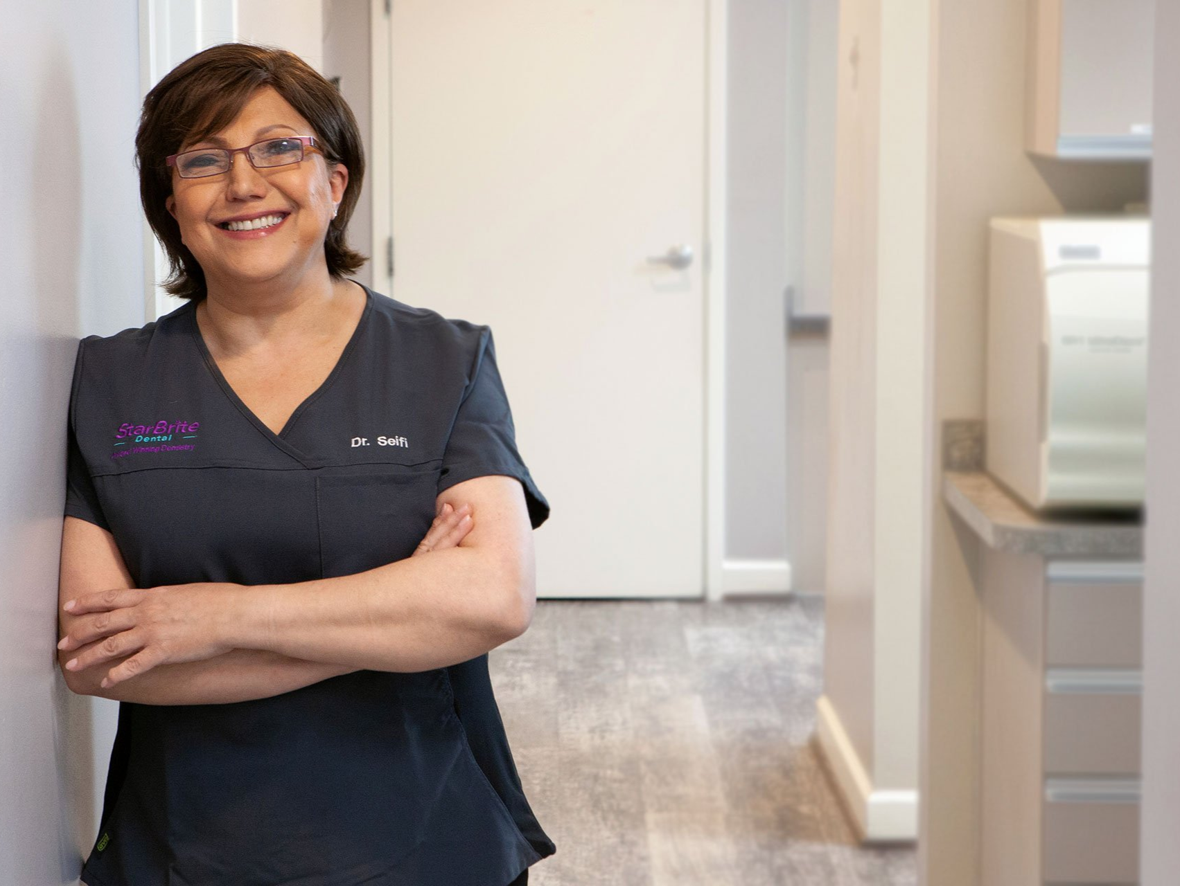 A woman is leaning against a wall with her arms crossed and smiling.