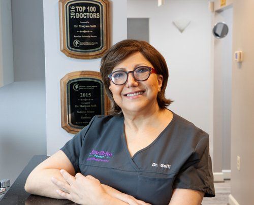 Dr. Seifi wearing scrubs and glasses is sitting at a desk in front of a wall with plaques on it.