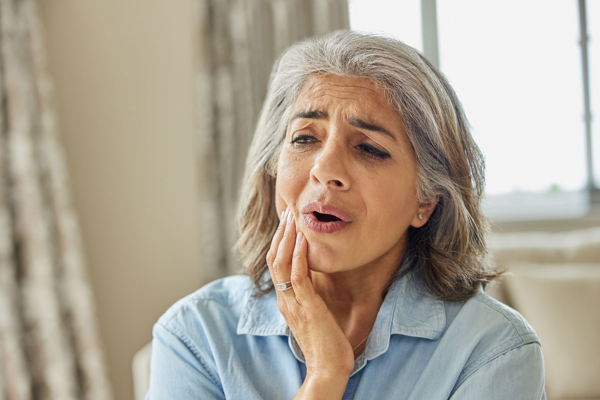 Woman with gray hair clutches her jaw, displaying an expression of pain, likely due to a toothache.