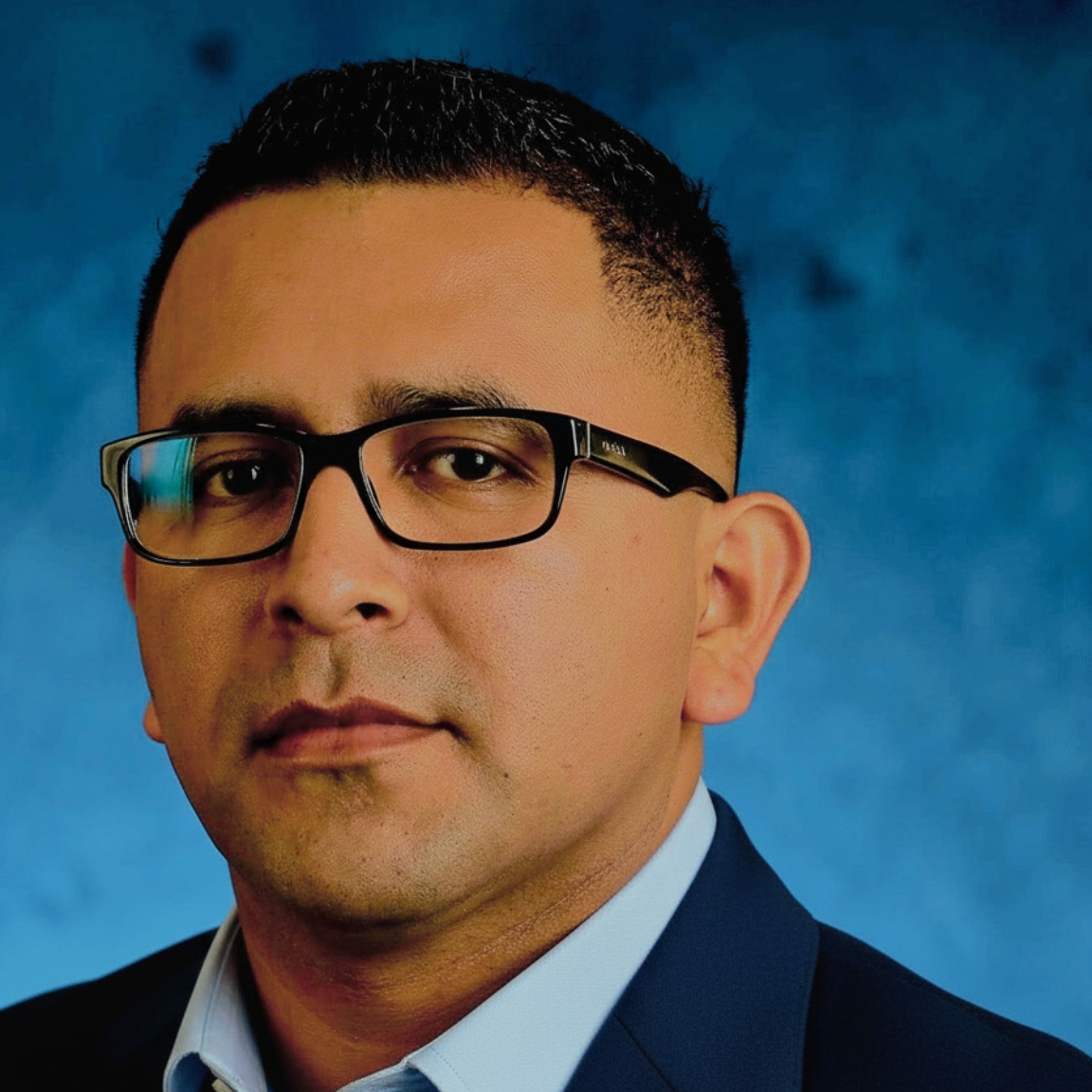 Man with glasses wearing a blue suit, looking forward, studio portrait with a blue background.