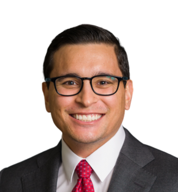 Man in suit and glasses smiles, red tie, against a blue patterned backdrop.