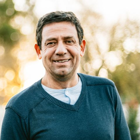 Man in blue sweater smiles outdoors, blurred background of trees and light.