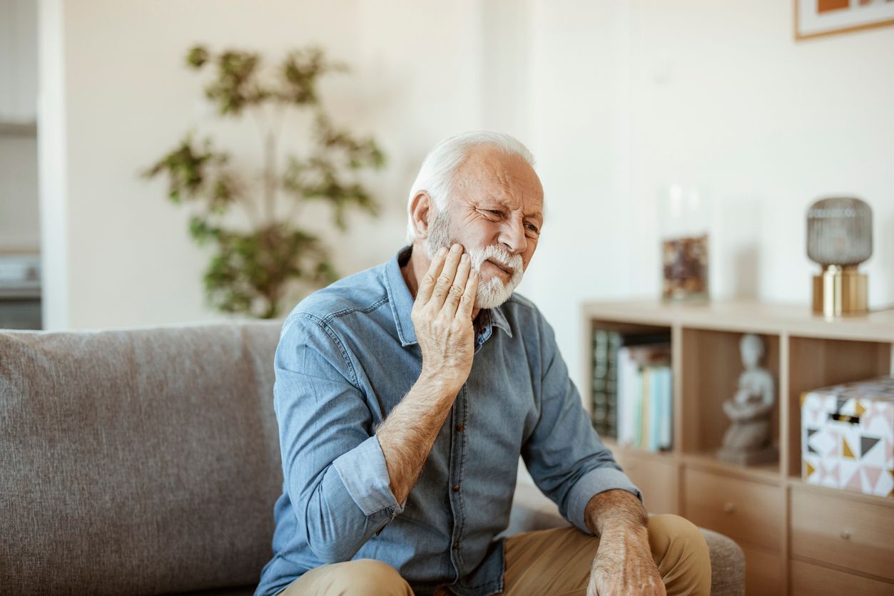 Man in blue shirt touching jaw, seated on sofa, appearing to be in pain.