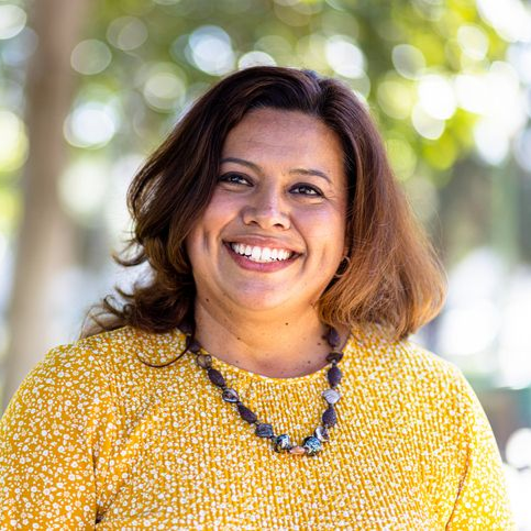 Woman with a warm smile, wearing a yellow sweater and necklace, outdoors with blurred greenery.