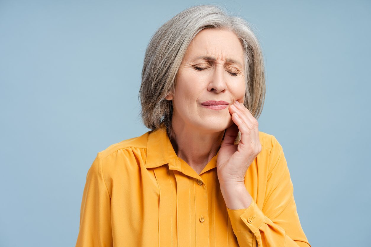 Woman with silver hair, eyes closed, holding her jaw in pain. Yellow shirt, blue background.