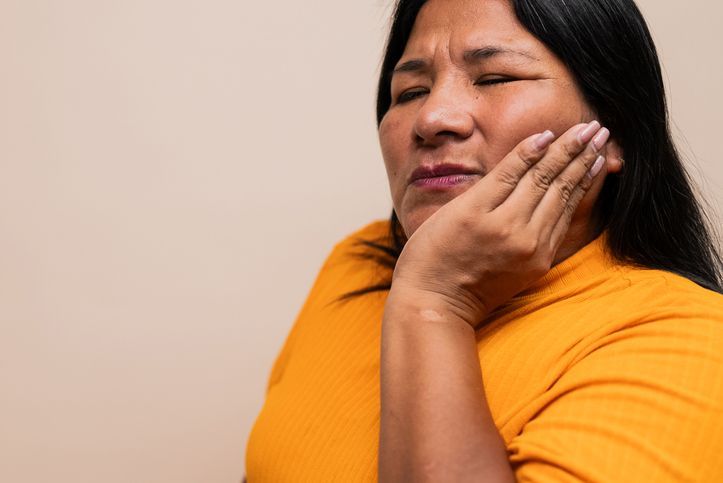 Woman with silver hair, eyes closed, holding her jaw in pain. Yellow shirt, blue background.