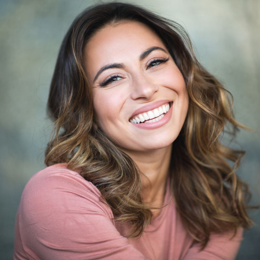 Woman with brown hair smiles, wearing a pink shirt, against a muted background.
