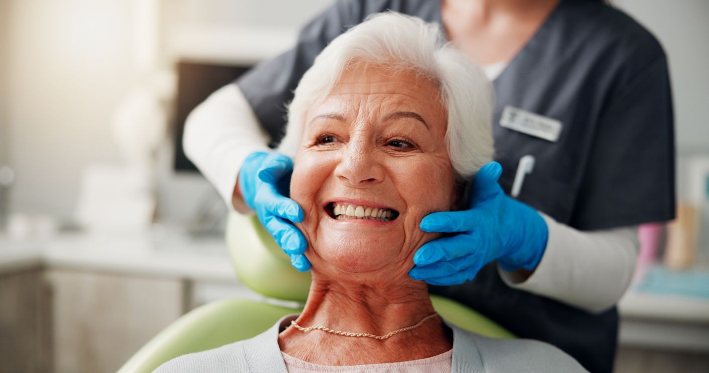 Dentist in blue gloves examining an elderly patient's mouth in a dental office. The patient smiles.
