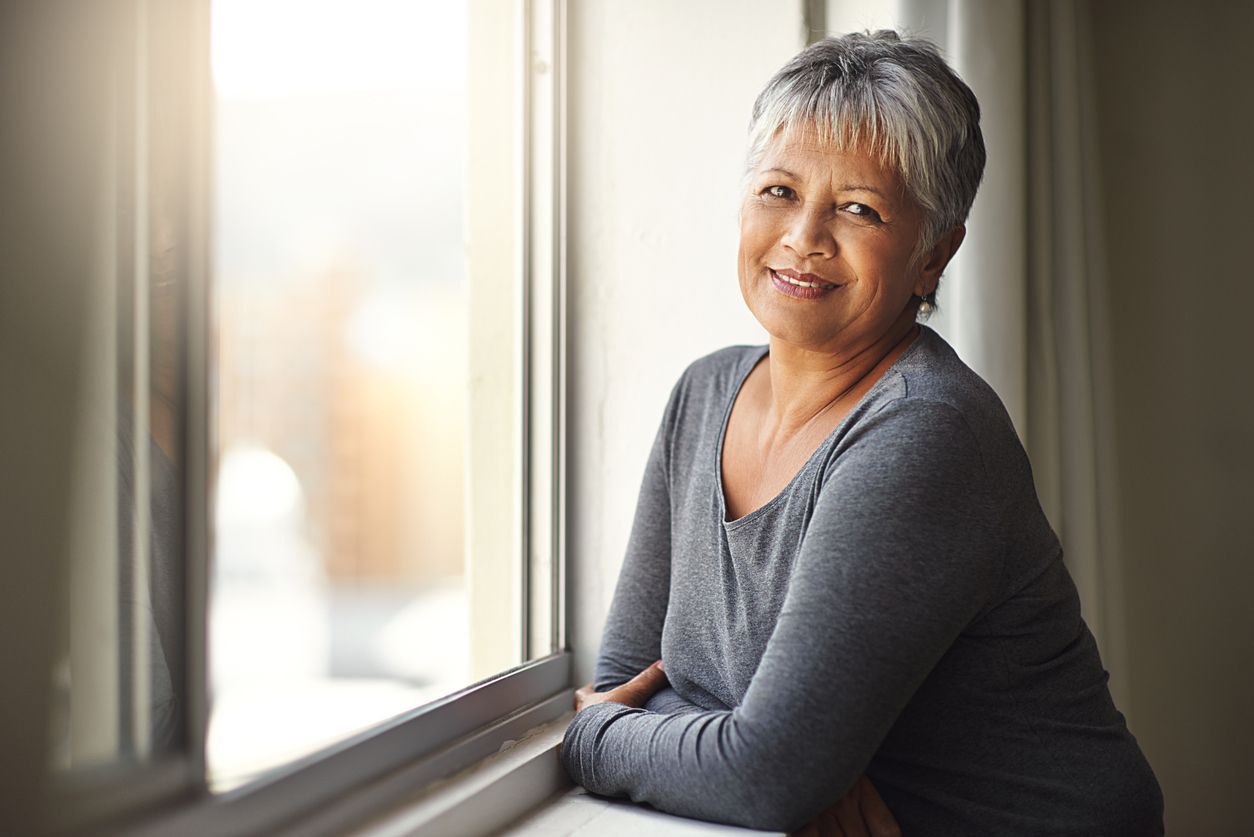 Woman with gray hair smiles, leaning on a window sill, looking at the camera.