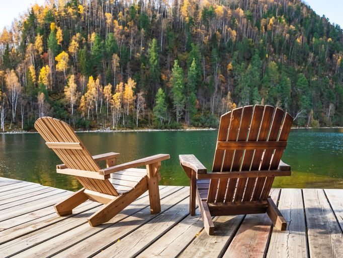 Two empty wooden Adirondack chairs or Muskoka deckchairs on wooden shore overlooking scenic calm Lake with mountains in the distance