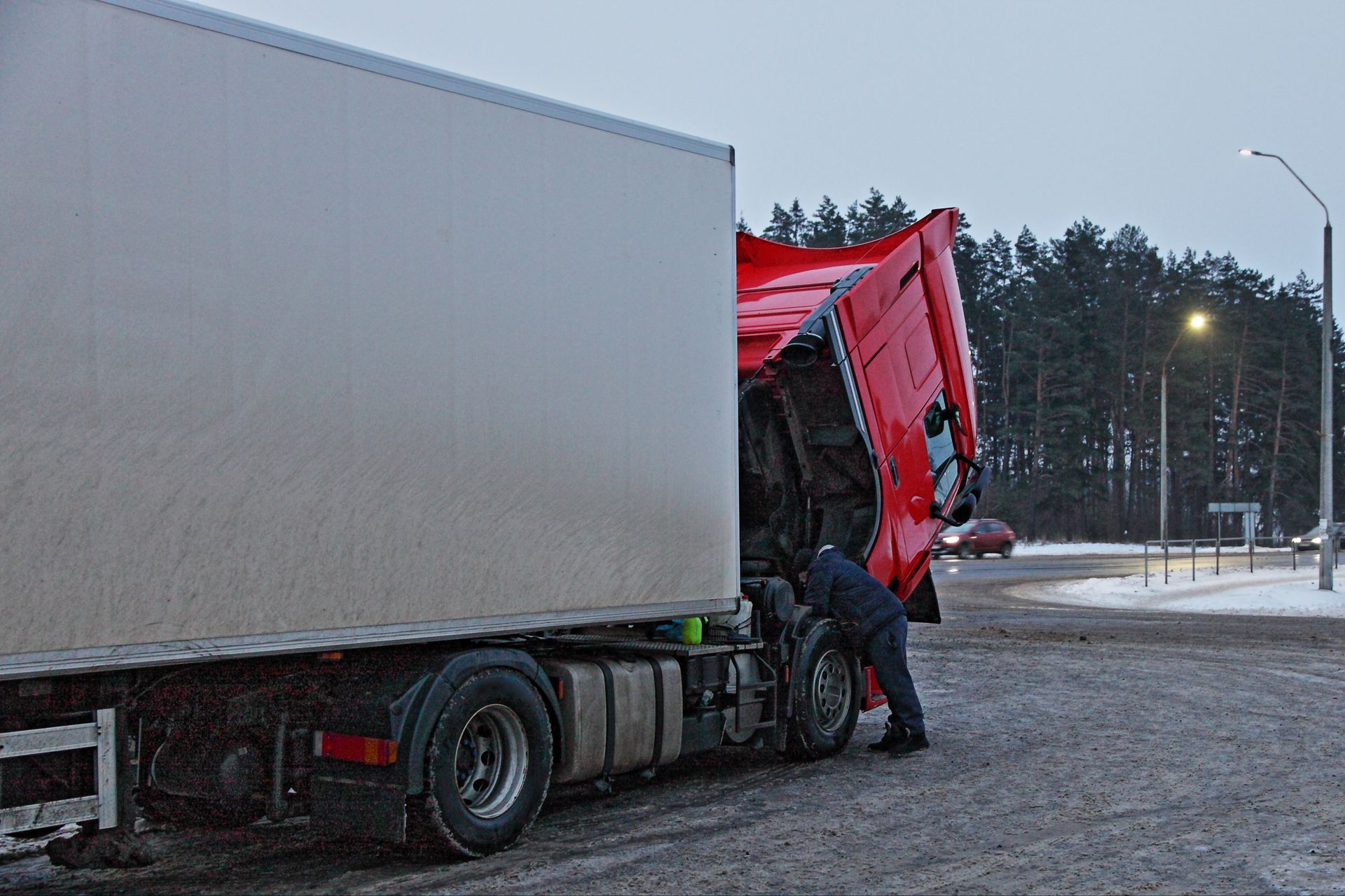 Mechanic inspecting the raised cab of a large trailer truck on a snowy roadside.