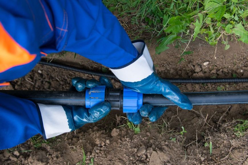 Hands wearing blue gloves connecting black pipes with a blue fitting in a dirt setting.