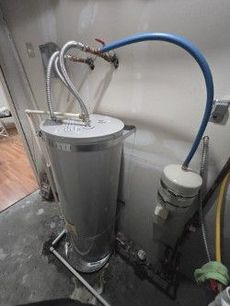 Gray water heater with flexible pipes and a blue hose in a concrete utility room.