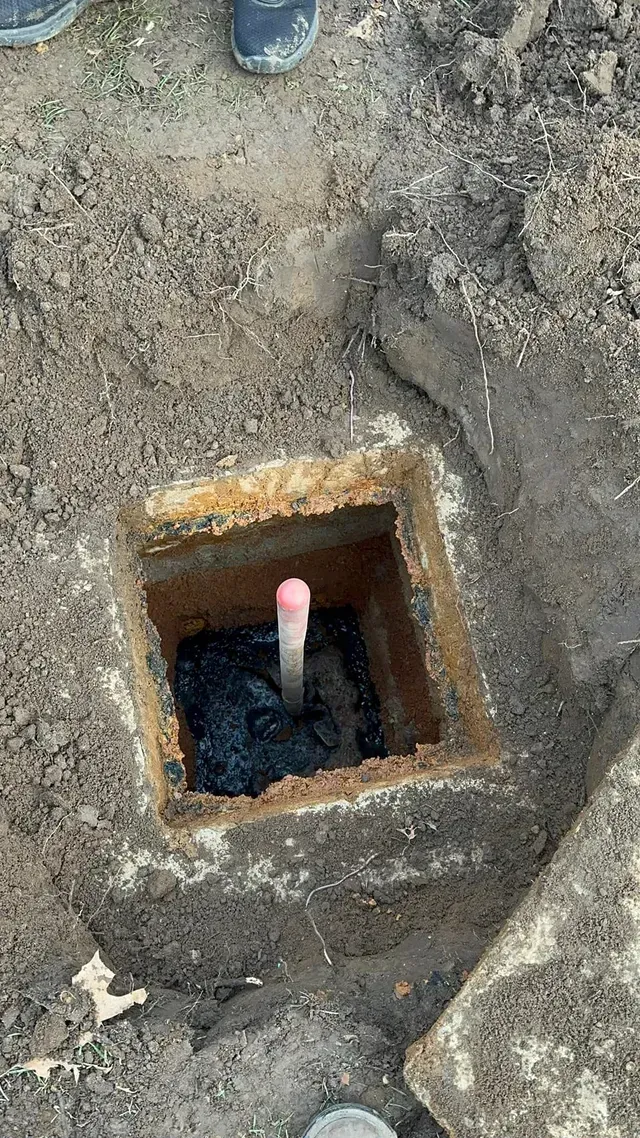 Square hole dug in dirt with a white pipe and dark material inside.