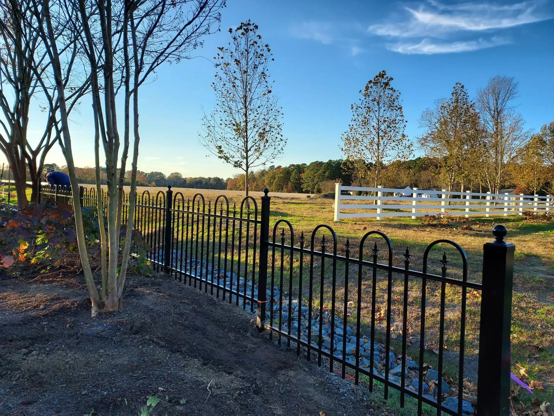 Black iron fence bordering a grassy field; white fence in the distance; blue sky.