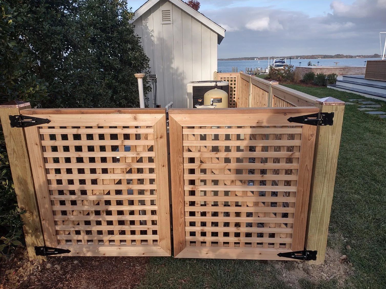 Double wooden lattice gate mounted on thick posts, concealing equipment near a coastal yard and small building.