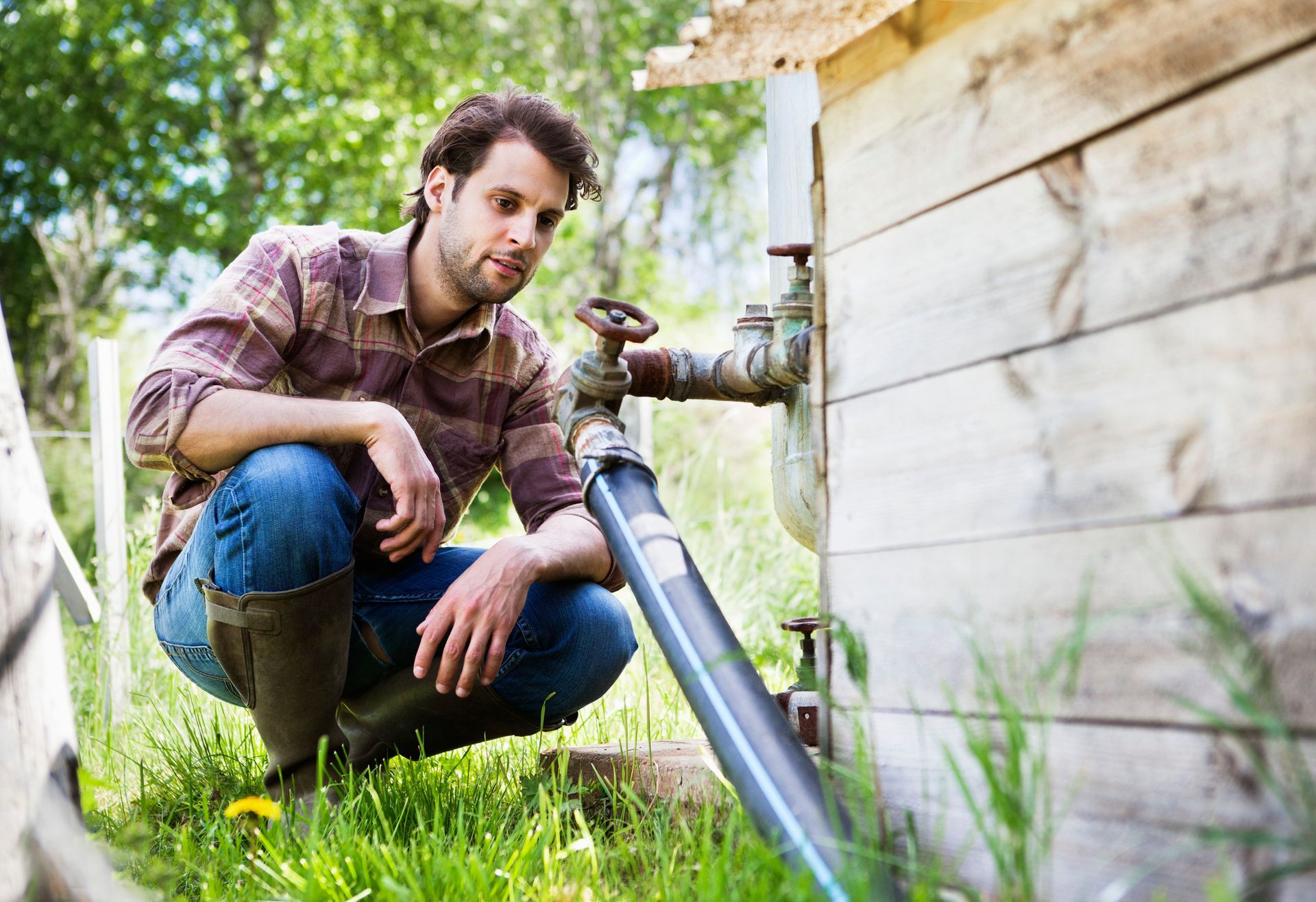 A man looking at a water pipe outdoors beside a wooden shed.