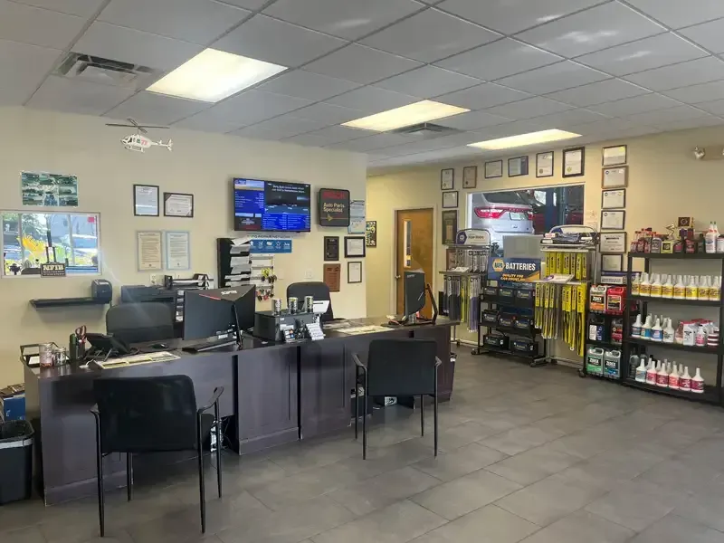 Interior of an auto repair shop's waiting area. Counters, computers, a wall of diplomas, and shelves of car fluids are visible.