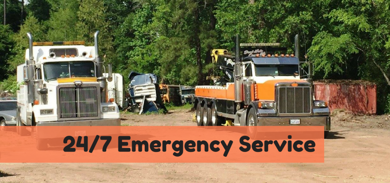 Two tow trucks with 24/7 Emergency Service banner. Trucks are white and orange, in a wooded area with a vehicle.