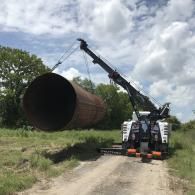 A crane lifting a large cylindrical metal object on a dirt road, cloudy sky in the background.