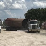 Truck hauling a large, cylindrical metal container in a lot with other equipment.