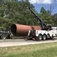 A large, cylindrical metal object being transported on a flatbed trailer, towed by a large tow truck, roadside.