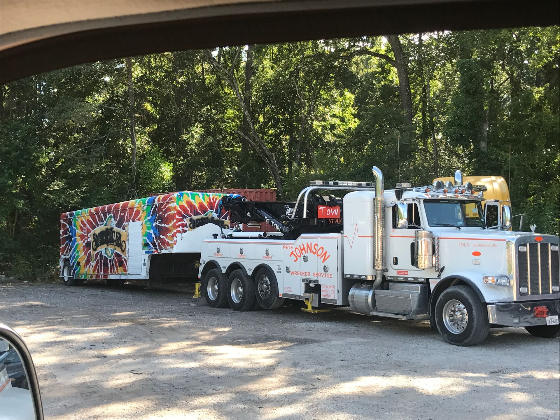 White tow truck towing a trailer with colorful tie-dye design; parked near trees.