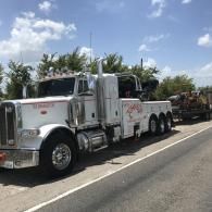 White tow truck hauling a trailer with debris on a road under a blue sky.