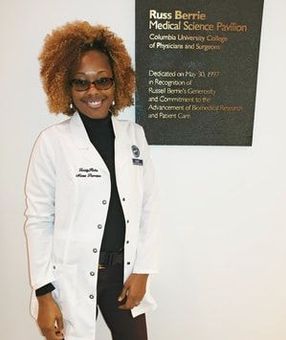 A woman in a white lab coat is standing in front of a sign.