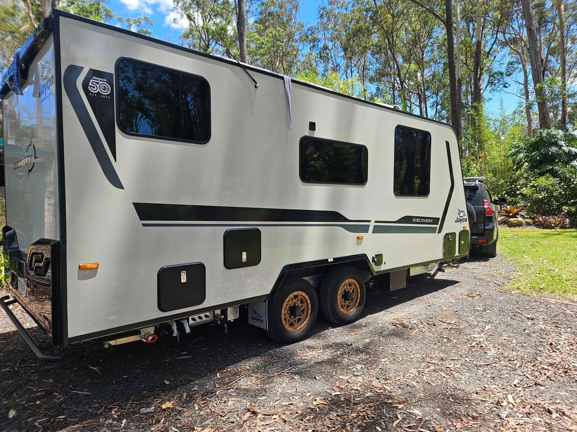 Gray and black caravan parked on gravel, with dark tinted windows and gold wheels, set in a grassy area.