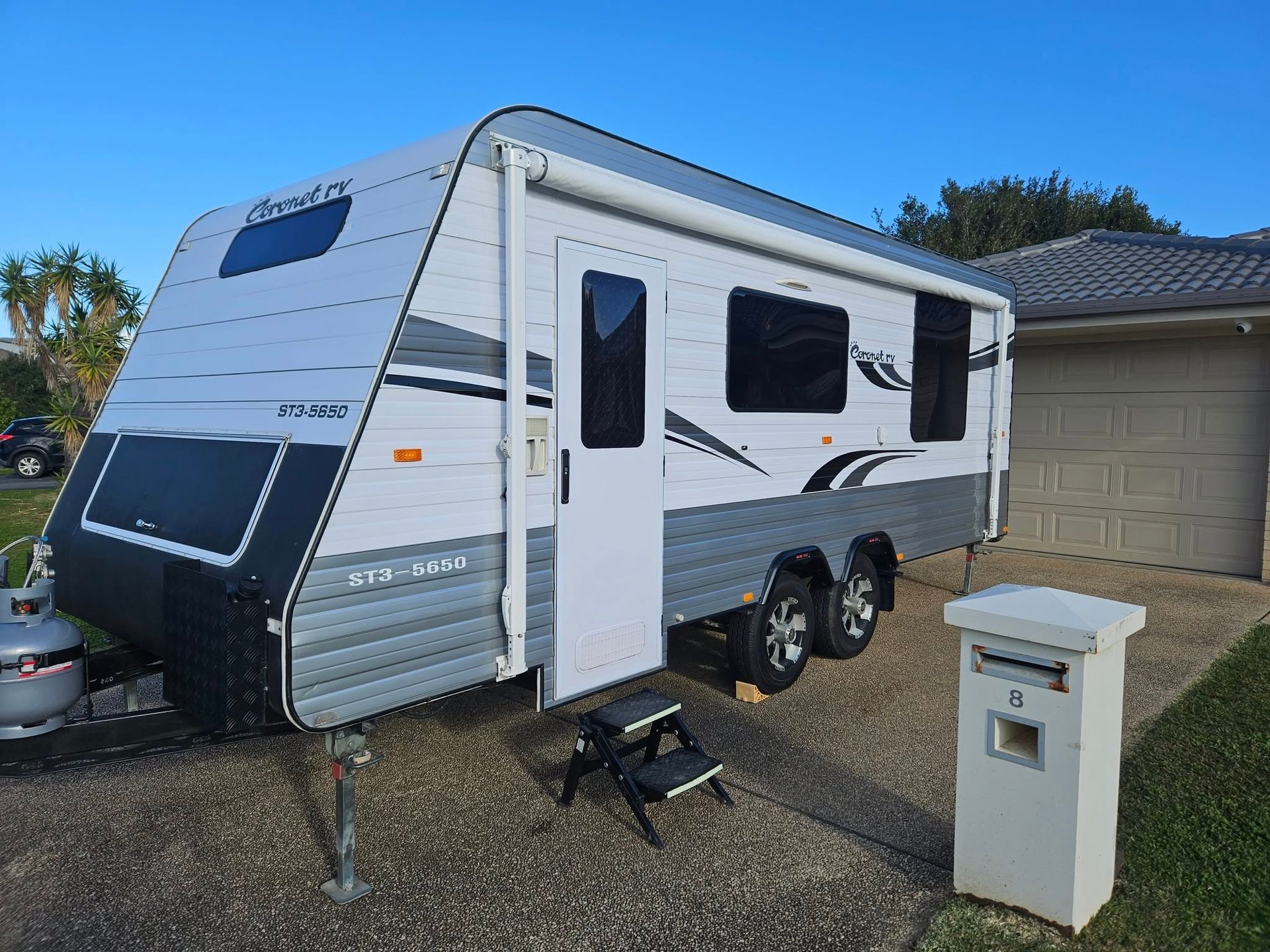 Gray and white caravan parked in front of a house, with a mailbox and steps in the foreground.