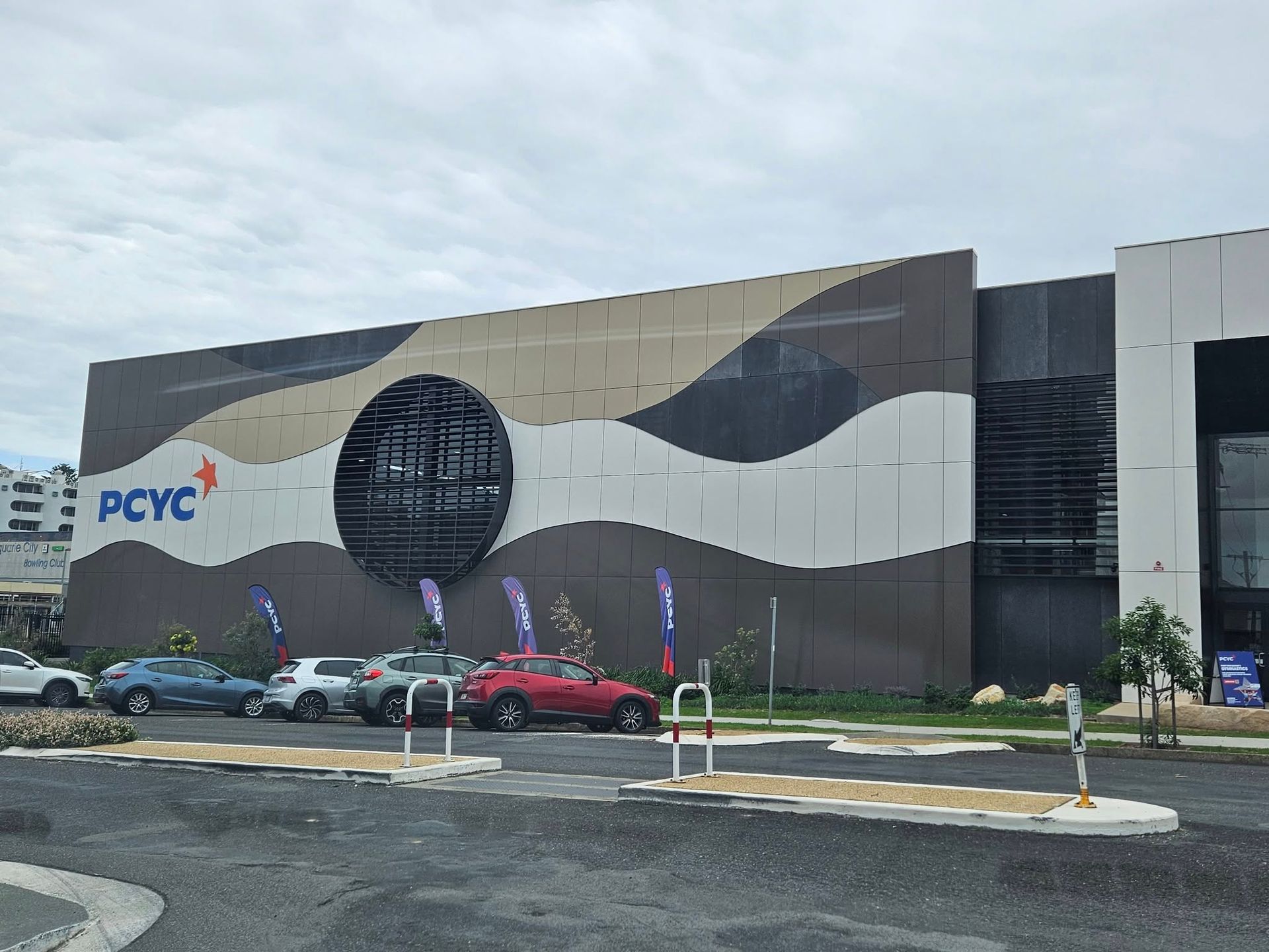 Exterior of PCYC building with wave-patterned facade, cars parked in front, cloudy sky.