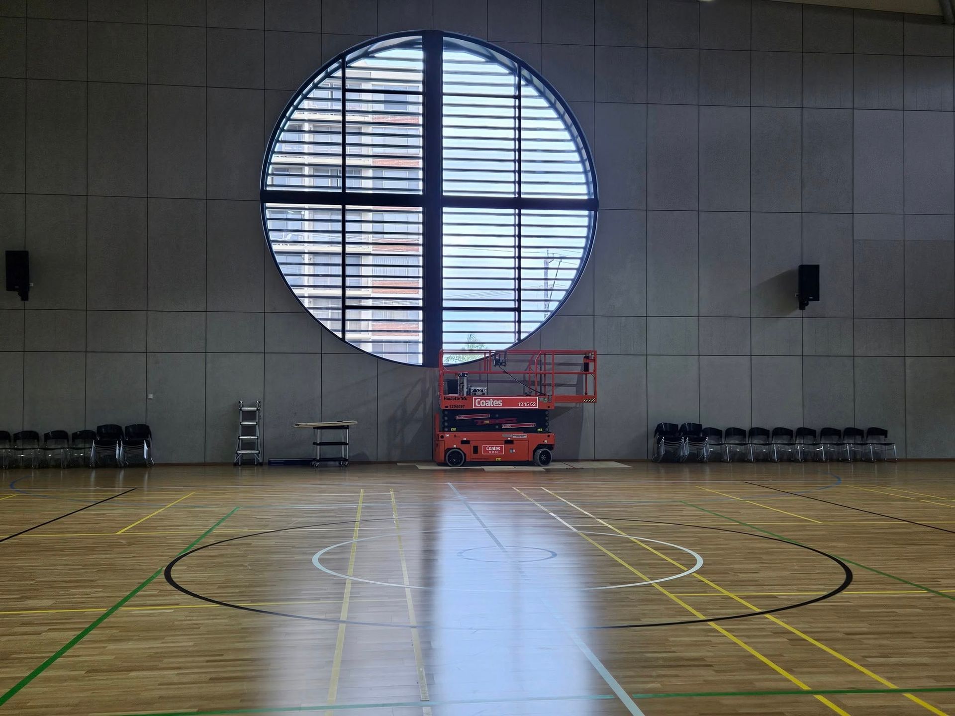 Indoor gymnasium with a large circular window, court markings, and maintenance lift.