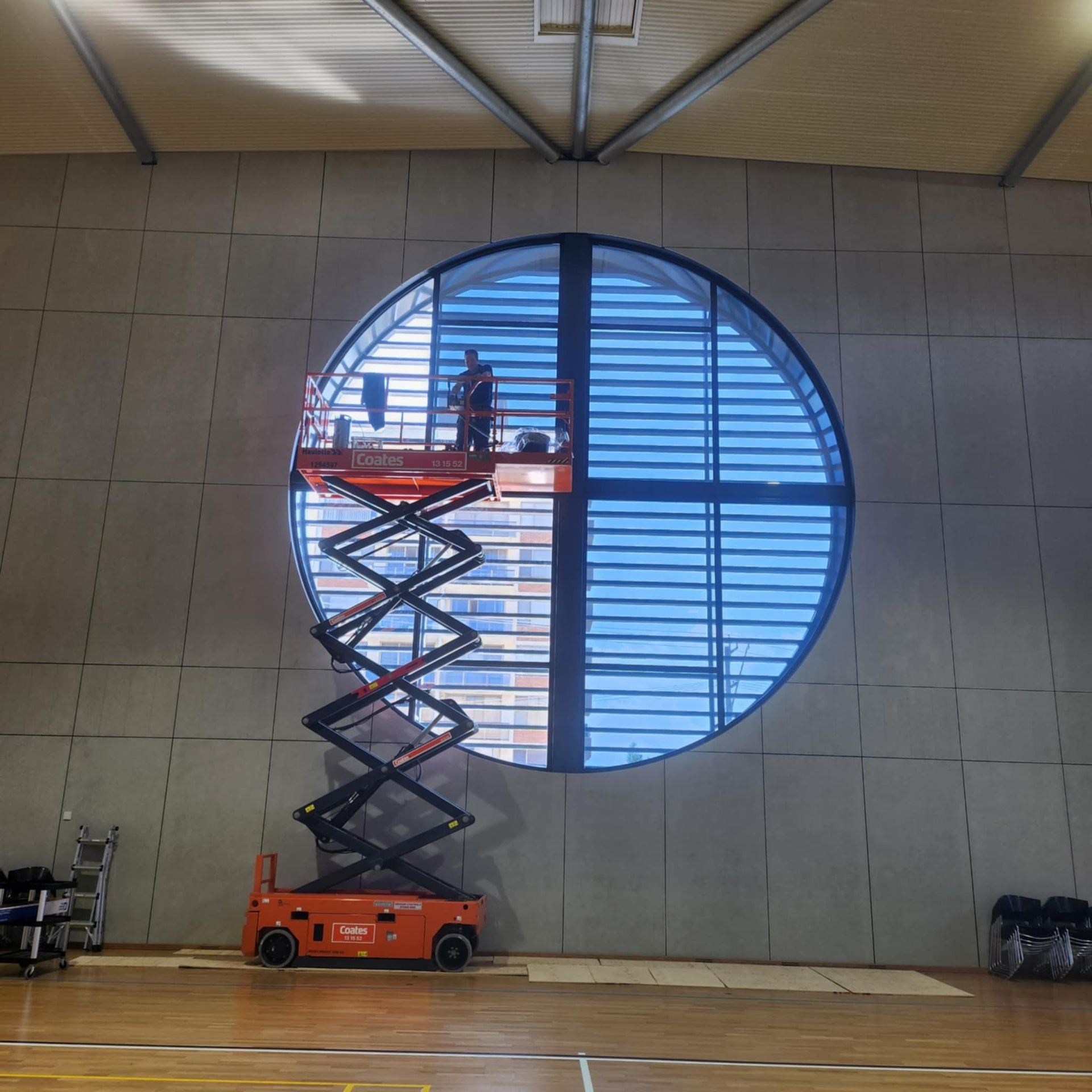 Person on a scissor lift working on a circular window in a gym. Buildings seen through the window.