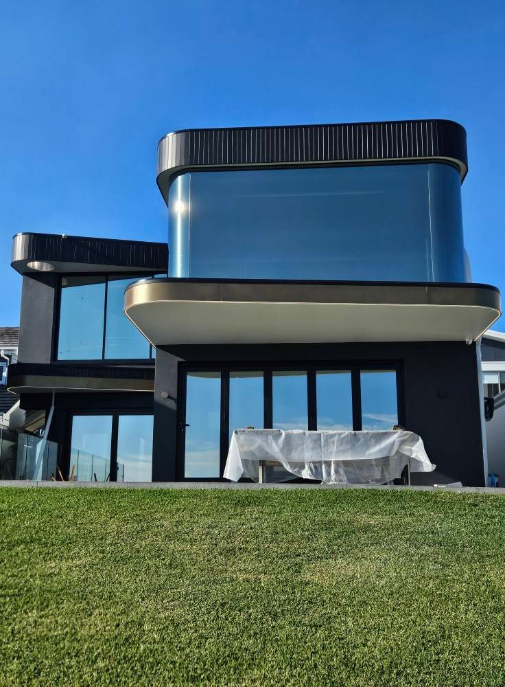 Modern black house with curved glass windows and gold trim against a blue sky, with a table covered on the patio.