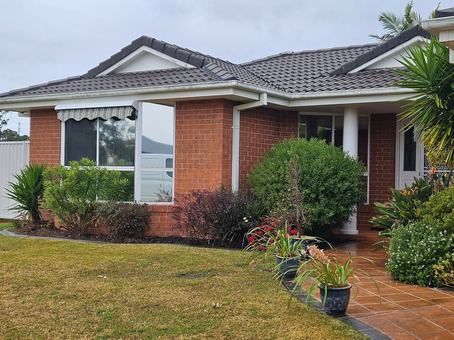 Brick house with dark roof, windows, and manicured landscaping, overcast sky.