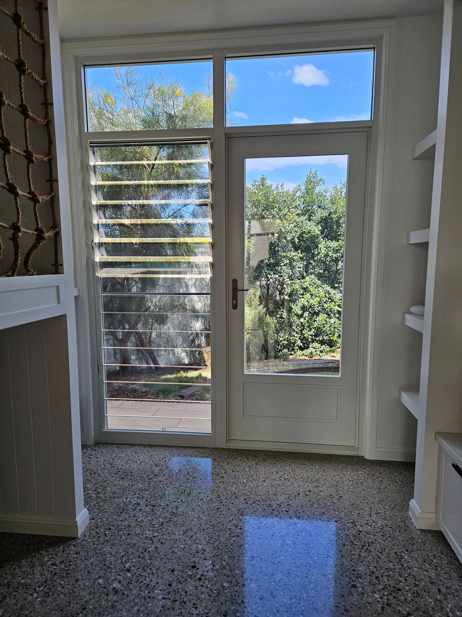 Doorway to outside: glass door with sunlight shining on a speckled floor. Outside, green trees and blue sky are visible.