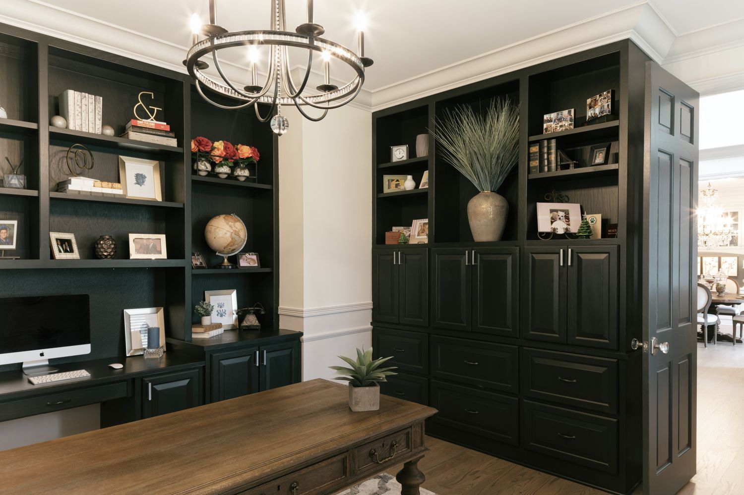 A home office with black cabinets , shelves , a desk and a chandelier.
