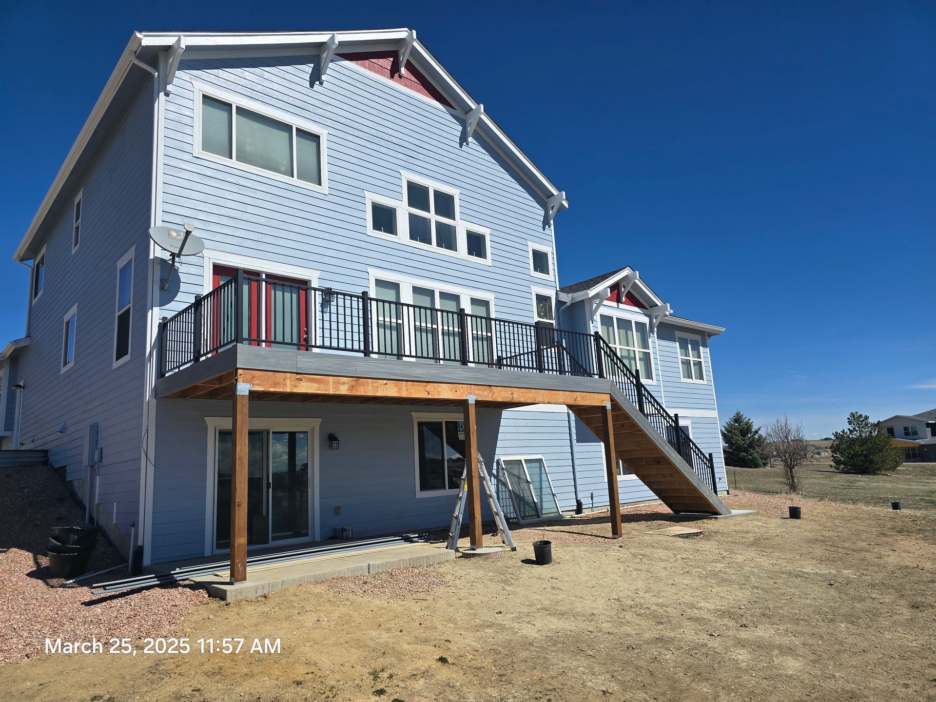 Blue two-story house under construction with a deck, stairs, and dry yard under clear sky