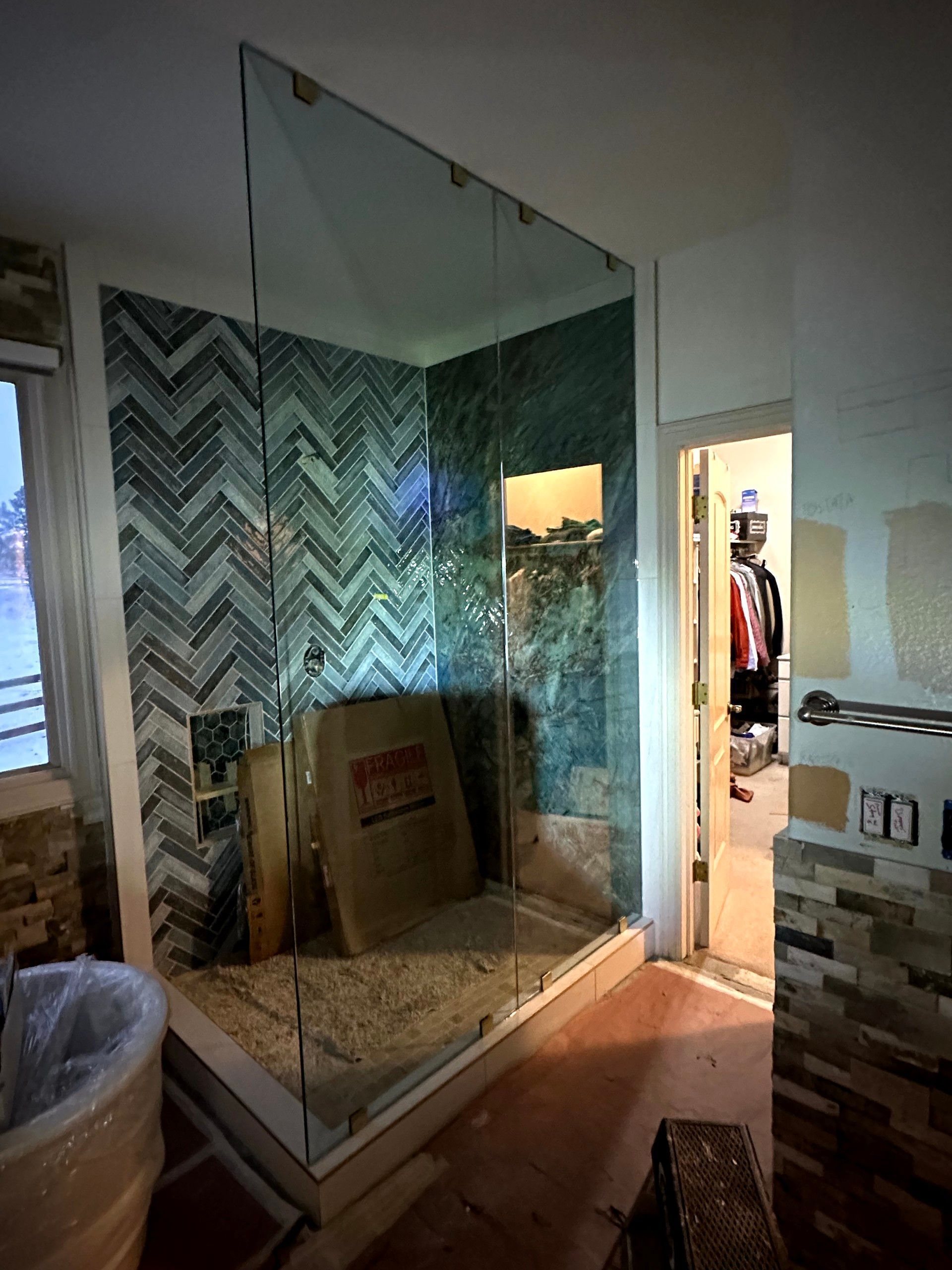 Glass shower enclosure under construction in a bathroom with herringbone tile and an open doorway.