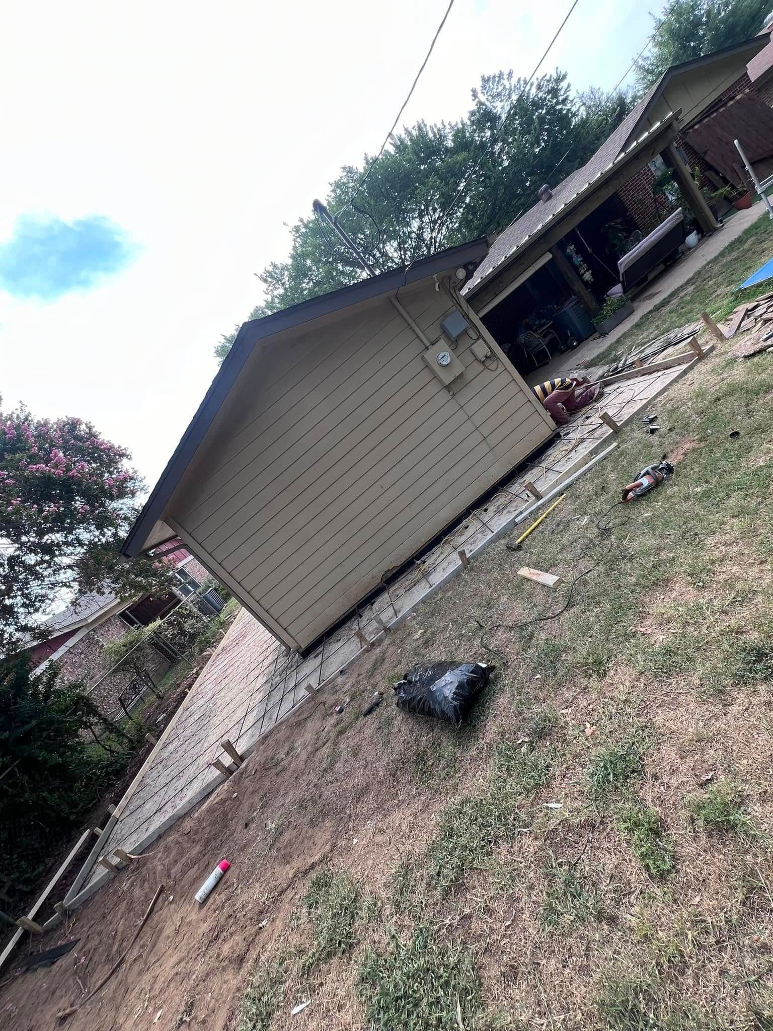 A shed on a sloped lawn with a partially visible house in the background. Blue sky and trees are present.