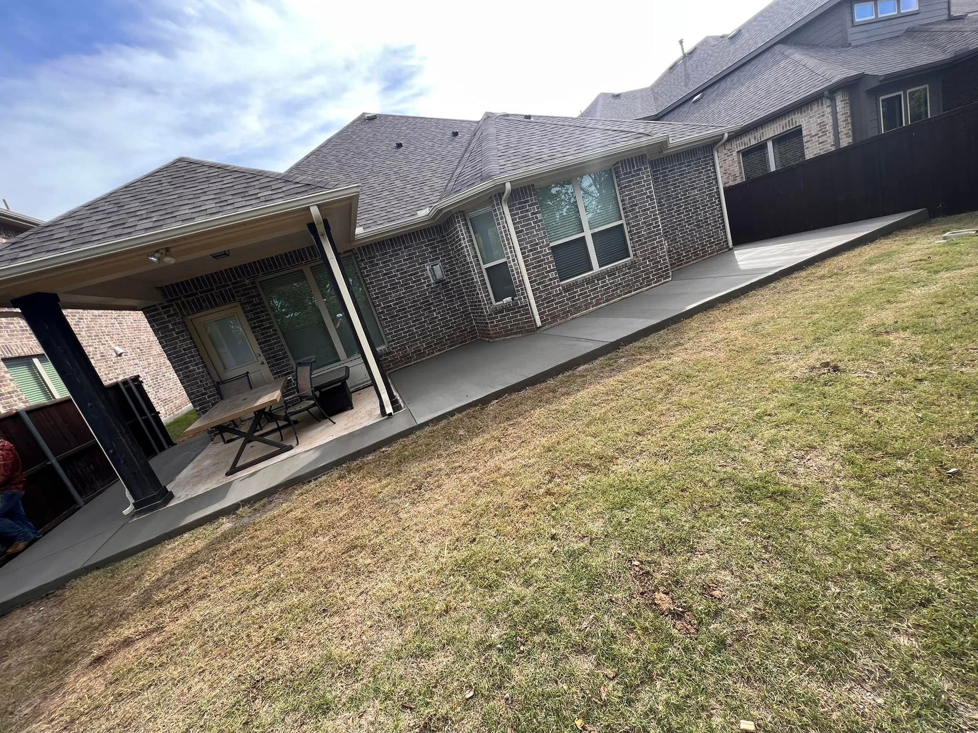 Backyard patio with covered seating area and house, on a slight slope, grass in foreground.