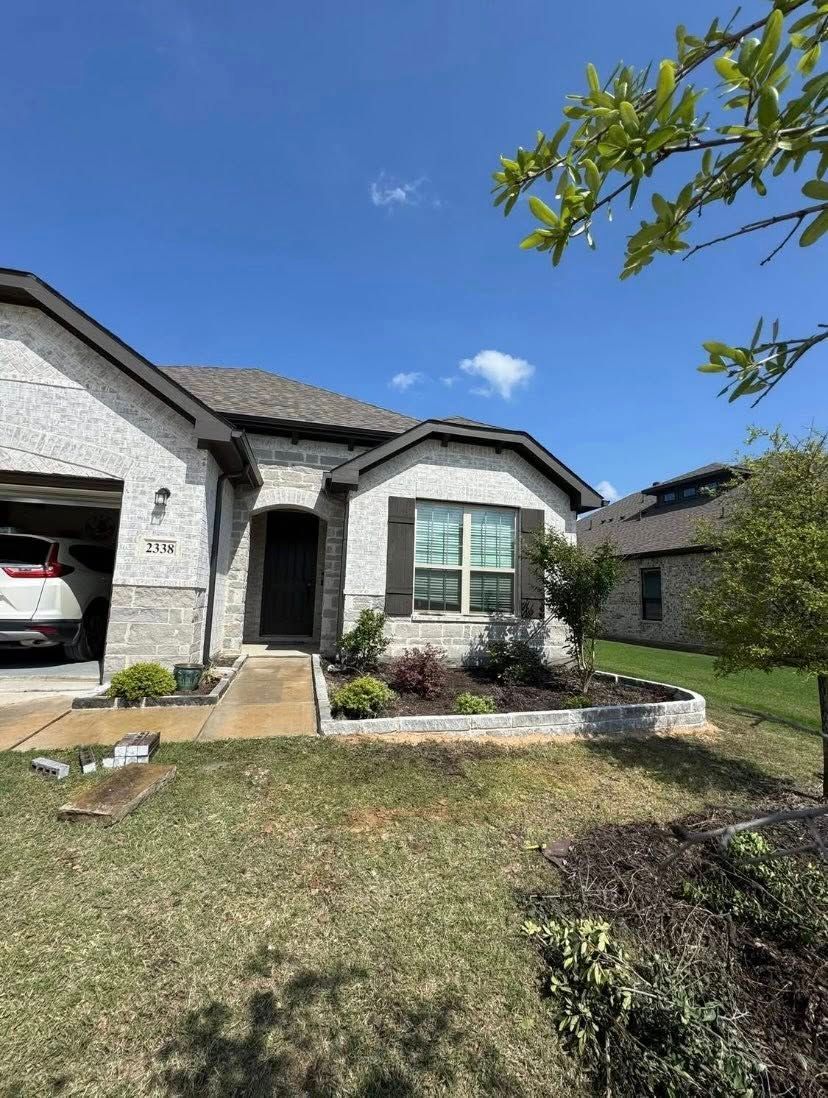 Light brick house with brown roof, front door, small landscaping, and driveway under a blue sky.