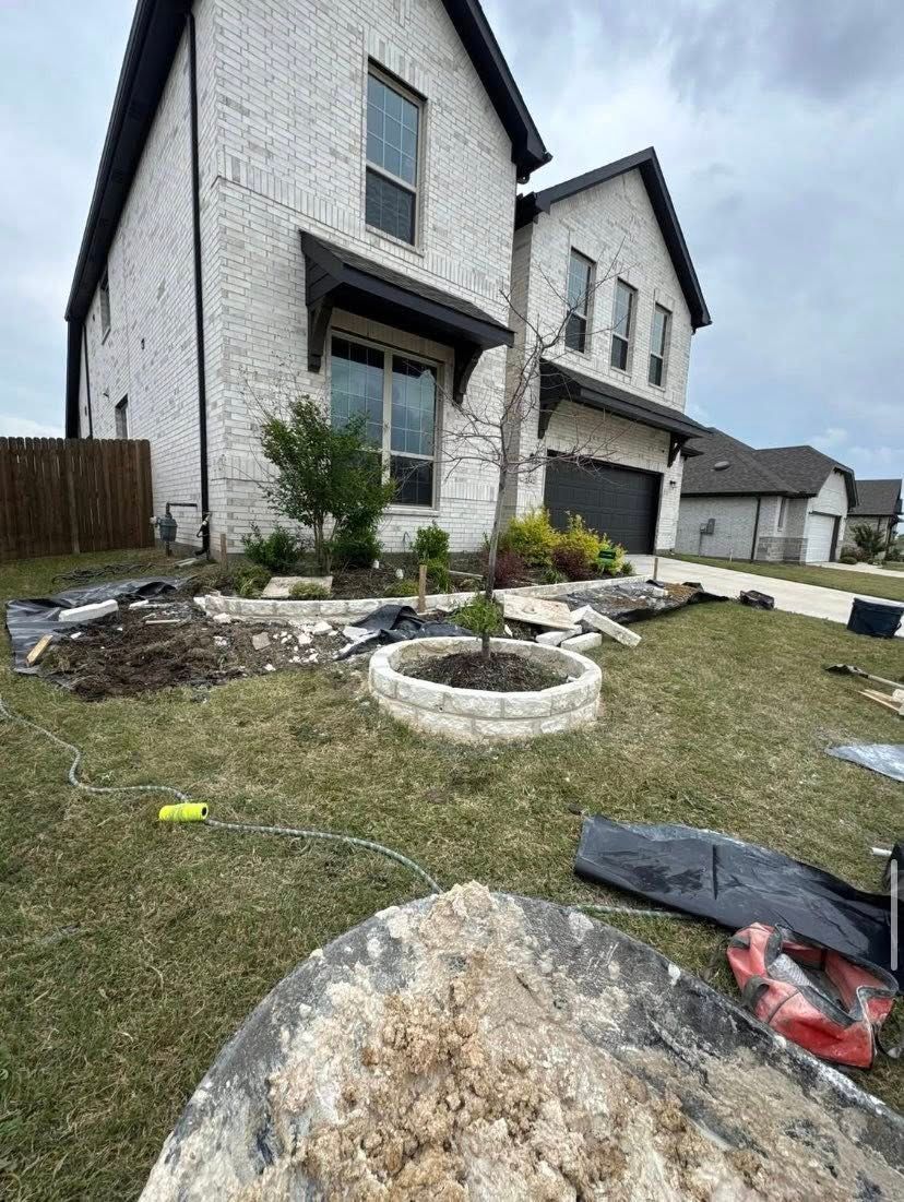 Exterior of a two-story brick home undergoing landscaping. A circular flower bed is visible in the yard.